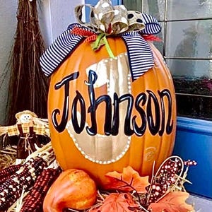 May include: An orange pumpkin decorated with the name "Johnson" in black script. The pumpkin is adorned with a decorative bow and sits on a bed of straw with fall-themed decorations, including corn and leaves.