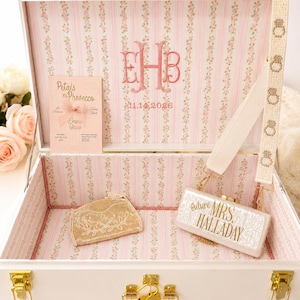 May include: A white keepsake box with gold latches, lined in pink floral striped fabric. Inside is a card reading Petals ni Prosecco, a gold beaded pouch, and a white clutch with the text future MRS. HALLADAY, featuring a strap with ring icons.