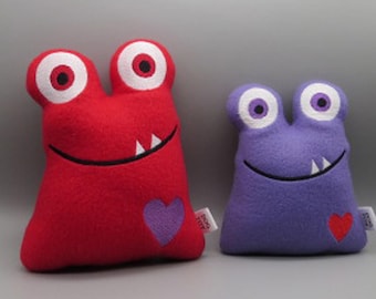 May include: Two plush toys shaped like monsters with big eyes and toothy grins. The larger monster is red with a purple heart, and the smaller monster is purple with a red heart.