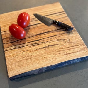 May include: A wooden chopping board with a natural wood grain pattern. Two red tomatoes and a black and silver knife are on the board. The board has a dark blue edge.
