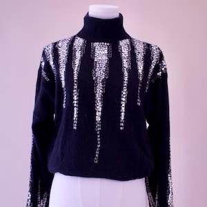 May include: A black turtleneck sweater with a silver sequin design. The sequins are arranged in a geometric pattern on the front and sleeves.