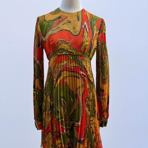 Pode incluir: Um vestido vintage com um padrão vibrante e rodopiante em tons de laranja, verde, amarelo e castanho. O vestido de manga comprida apresenta uma saia plissada e uma cintura ajustada. O vestido é exibido em um manequim contra um fundo liso.