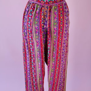Può includere: Pantaloni a vita alta e fantasia con un vivace mix di colori. I pantaloni presentano una base rossa con strisce verticali di vari motivi in viola, verde, giallo e blu. La cintura presenta un design colorato e dettagliato.