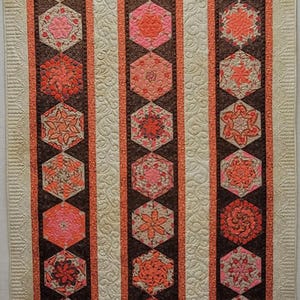 May include: A quilt with three vertical rows of orange and brown hexagonal patterned fabric blocks. The blocks are arranged in a repeating pattern. The background fabric is a light beige with a subtle floral pattern.