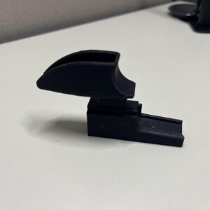 K&ouml;nnte beinhalten: Ein mattschwarzes, 3D-gedrucktes Kunststoffbauteil mit einem geschwungenen, hohlen Oberteil, das an einer rechteckigen Basis mit einer gerillten, l&auml;nglichen Verl&auml;ngerung befestigt ist. Das Objekt hat eine starre, geometrische Struktur.