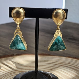 May include: A pair of gold earrings with green gemstone triangle drops. The earrings have a textured gold finish and are on a black stand.