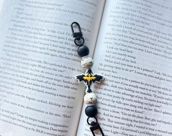 May include: A black and white beaded bookmark with a black metal clip and a black and yellow bat charm. The beads are round and have a variety of patterns, including polka dots, stripes, and a skull and crossbones. The bookmark is lying on an open book with text.