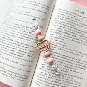 Puede incluir: Un llavero en forma de corazón rosa y marrón con un amuleto de bandeja de comida de madera en miniatura. La bandeja presenta una variedad de artículos de comida en miniatura de colores, incluyendo una rebanada de sandía, una fresa, un cupcake y una rebanada de pizza. El llavero también presenta un amuleto de cuentas rosa y blanco.