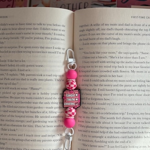 Może przedstawiać: Różowo-biały brelok z koralików ze srebrnym zapięciem, z zawieszką z napisem "Single Taken Reading". Brelok jest przymocowany do stron książki.