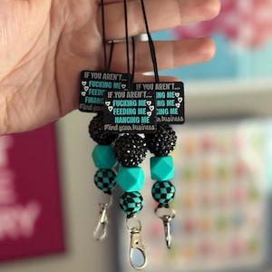 May include: Three lanyards featuring black rectangular tags with white and teal text reading IF YOU AREN'T... FUCKING ME, FEEDING ME, FINANCING ME, Mind your business. Each includes black glittery beads, teal geometric beads, and black and teal checkered beads.