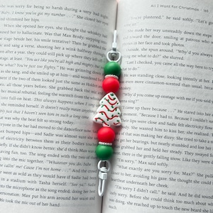 May include: A Christmas-themed bookmark with a silver-tone metal clip. The bookmark features green and red beads, and a white tree-shaped charm with red and green accents. The bookmark is placed in an open book.