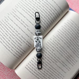 May include: A beaded book charm with a black clasp, black and white beads, and a white rectangular charm with the text "JUST ONE MORE CHAPTER" and a skull graphic. The charm hangs over an open book.