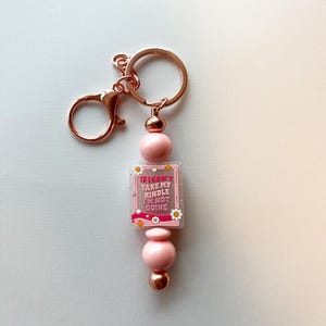 Op de afbeelding: Een roze en roségouden sleutelhanger met een roze vierkante hanger waarop staat "Ik kan mijn Kindle niet meenemen, ik ga niet".
