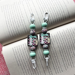 May include: Two colourful book-themed keychains with black and white patterned beads, a green charm, and a silver clasp. The charms feature a stack of books with the words "Books On" and "Books Off" on them.