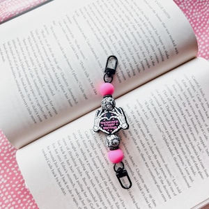 May include: A pink and black charm with a heart-shaped design and the text "You Had Me At Trigger Warnings". The charm is attached to a black and silver chain with pink beads.