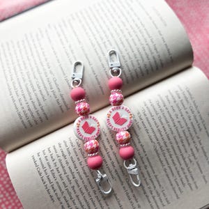 May include: Two pink and white beaded keychains with a book charm. The charms have the text "A Book A Day Keeps Reality Away".