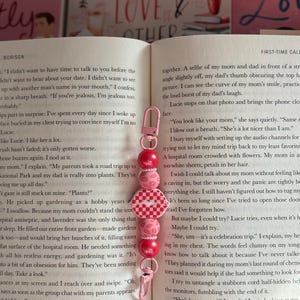 May include: Pink and red beaded keychain with a lobster clasp. The keychain features a series of round beads and a charm with a checkered pattern and a lip design. The keychain is shown on top of an open book.