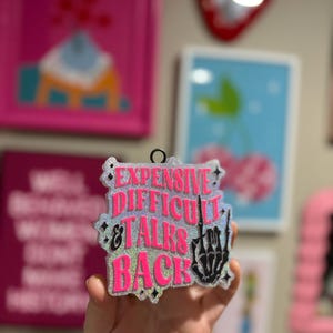 May include: A pink and silver glitter bauble with the text "Expensive Difficult & Talks Back" and a black skeleton hand giving the middle finger.
