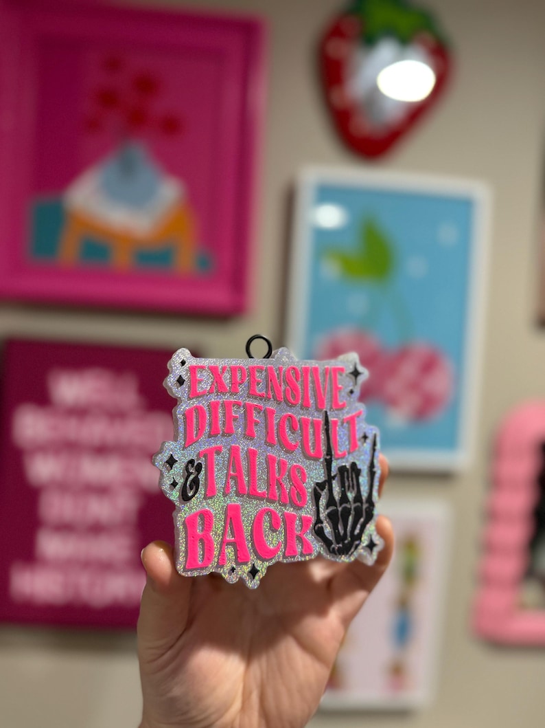 May include: A pink and silver glitter bauble with the text "Expensive Difficult & Talks Back" and a black skeleton hand giving the middle finger.