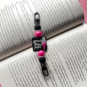 May include: A black and pink beaded bookmark with a skeleton hand holding a book and the text "Spread those pages".