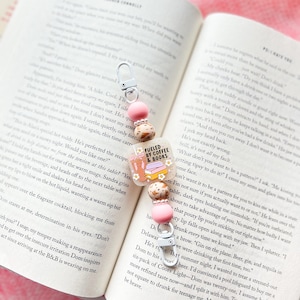 May include: A beaded bookmark with a charm that reads "Fueled by Coffee & Books." The bookmark features pink beads, small brown beads, and a silver clasp. The charm has a coffee cup and book design.