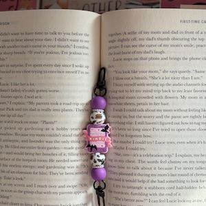 May include: A purple beaded Kindle charm with a black clasp hangs over an open book. The charm features a pink Kindle-shaped accent with bats and the text "A Reader Without Kindle".