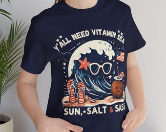 May include: Navy blue t-shirt with a graphic of a wave, sunglasses, starfish, and a coconut drink. The text reads "Y'all Need Vitamin Sea" and "Prescribed: Sun, Salt & Sass". The design includes red, white, and blue elements.