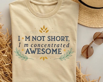 May include: A beige t-shirt with the text "I'M NOT SHORT, I'm concentrated AWESOME" in navy blue and gold. The design includes decorative floral elements. A straw hat and sunglasses are in the background.