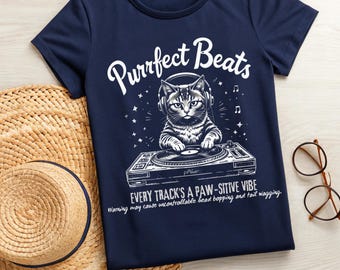 May include: A navy blue t-shirt featuring a white graphic of a cat wearing headphones and mixing music on a turntable. The text "Purrfect Beats" is above the cat and "Every track's a paw-sitive vibe" is below the cat. The text "Warning may cause uncontrollable head bopping and tail wagging." is at the bottom of the graphic.