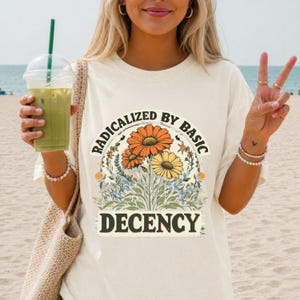 May include: A cream-colored t-shirt featuring a graphic print of orange and yellow daisies with blue wildflowers. The text RADICALIZED BY BASIC DECENCY is printed in black, arching over the flowers and centered below them in a bold font.