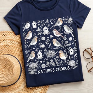 May include: Navy blue t-shirt featuring a detailed illustration of various birds, birdhouses, and floral elements. The design includes the text "NATURE'S CHORUS" in white. The shirt is a crew neck style and appears to be made of a soft fabric.