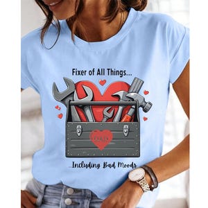 May include: Light blue t-shirt with a graphic featuring a toolbox, tools, and a red heart. The text reads "Fixer of All Things... Including Bad Moods." The word "DAD" is on the toolbox.