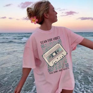 以下が含まれることがあります： カセットテープのグラフィックと「SCAN FOR VIBES」と「THIS SHIRT HAS SECRETS」の文字が入ったピンク色のTシャツ。ビーチで人が着ています。
