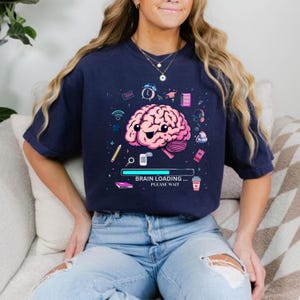 May include: Navy blue t-shirt with a pink cartoon brain graphic, smiling and surrounded by icons. The text "BRAIN LOADING... PLEASE WAIT" is below the brain. The shirt is a casual style, suitable for everyday wear.