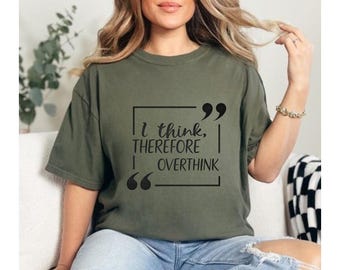 May include: Olive green t-shirt with the quote "I think, therefore overthink" in black text within a black box. The shirt is a relaxed fit and has short sleeves. The model is wearing blue jeans.