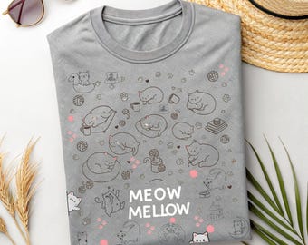 May include: A gray t-shirt with a repeating pattern of cartoon cats in various poses, along with paw prints and other cat-related illustrations. The words "MEOW MELLOW" are printed in white. The shirt is folded on a white surface.