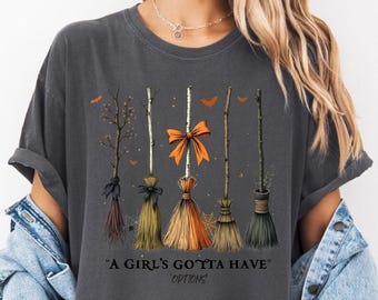 Könnte beinhalten: Dunkelgraues T-Shirt mit einem Design von fünf Besen mit orangefarbenen Schleifen und Akzenten. Der Text "A Girl's Gotta Have Options" ist darunter gedruckt. Das T-Shirt hat eine lockere Passform.