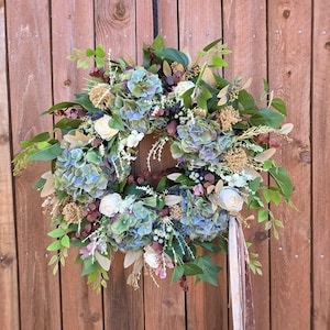 May include: A circular wreath featuring blue and green hydrangea blossoms, white roses, and various green foliage. The arrangement includes small tan floral accents and dark berries, finished with a long, light-colored ribbon hanging from the bottom.