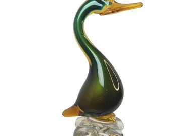 Peut inclure: Une figurine de canard en verre avec un corps vert profond à la finition lisse et réfléchissante. La tête et le bec sont en verre ambré translucide. Le canard repose sur une base en verre transparent texturé avec des bulles d'air visibles.