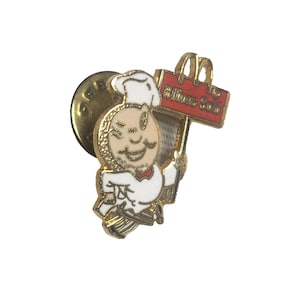Puede incluir: Un pin de solapa de metal dorado que muestra un personaje de chef de dibujos animados con uniforme y gorro blancos, sosteniendo un letrero rectangular rojo con el texto Billions Sold y un arco dorado de McDonald's, con un cierre circular.