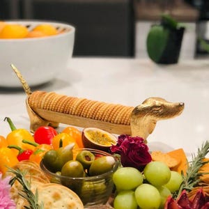 May include: A gold-toned metal dachshund-shaped cracker holder featuring a long, narrow body designed to hold a row of round crackers, with a detailed dog head at one end and a tail at the other, positioned among various fresh food items.