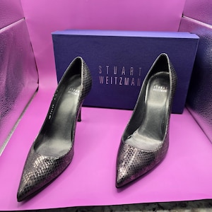 Può includere: Un paio di décolleté nere in pelle di serpente con punta a punta e tacco a spillo. Le scarpe sono su una superficie rosa e hanno il testo "Stuart Weitzman" sulla scatola.