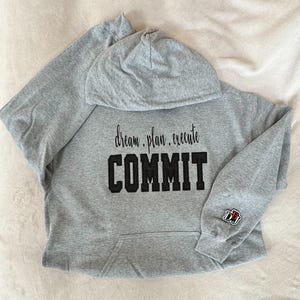 May include: Gray hooded sweatshirt with the text "dream. plan. execute COMMIT" in black lettering. The sweatshirt has a small black patch on the sleeve with the letters "DJ" in white.