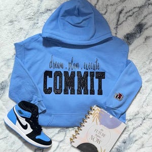 May include: A pale blue hoodie featuring the text "dream. plan. execute COMMIT". A pair of blue, black, and white trainers and a notebook with the words "the future begins NOW" are also visible in the image.
