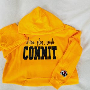 May include: Yellow hooded sweatshirt with black text that reads "dream. plan. execute COMMIT". The sweatshirt has a small black and red logo on the right sleeve.