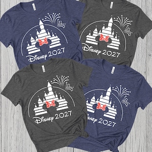 May include: Four short-sleeved t-shirts in navy blue and gray, each featuring a white castle design with a red bow and the text "Disney 2027". The shirts are arranged on a light wood-grain surface.