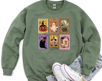 May include: Green sweatshirt featuring Halloween-themed postage stamp designs. The stamps depict a jack-o'-lantern, ghost, cauldron, black cat, haunted house, and skull with the text "Trick or Treat". The sweatshirt is paired with denim shorts and white sneakers.