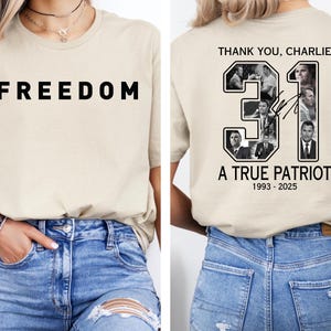 K&ouml;nnte beinhalten: Beiges T-Shirt mit dem Wort "FREEDOM" in gro&szlig;en schwarzen Buchstaben auf der Vorderseite. Auf der R&uuml;ckseite des Shirts befindet sich ein Design mit dem Text "THANK YOU, CHARLIE", der Zahl "31" und den Worten "A TRUE PATRIOT 1993-2025".