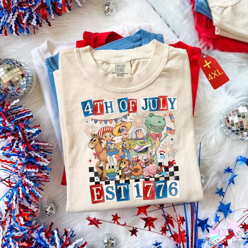 May include: A stack of t-shirts in white, blue, and red, with a cream-coloured shirt on top featuring a graphic for the 4th of July. The graphic includes cartoon characters and the text "4th of July EST 1776". A red tag reads "4XL".