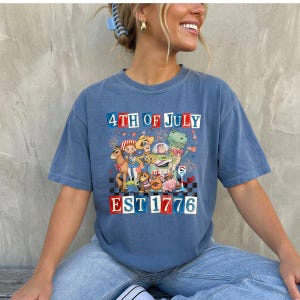 May include: Blue t-shirt with a graphic featuring cartoon characters and the text "4TH OF JULY EST 1776" in red, white, and blue. The shirt has a vintage, faded appearance.